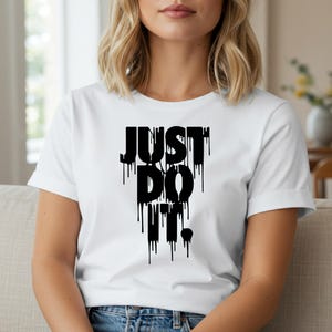Puede incluir: Camiseta blanca con el texto negro "JUST DO IT" en una fuente goteante. La camiseta tiene mangas cortas y cuello redondo. La modelo lleva la camiseta.