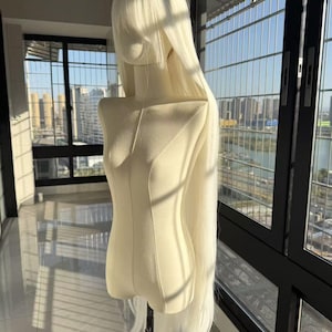 Puede incluir: Un maniquí de cabeza y torso con una larga peluca blanca. La peluca tiene cabello liso y fluido que se extiende más allá de la cintura del maniquí. El maniquí es de color crema y está frente a una ventana.