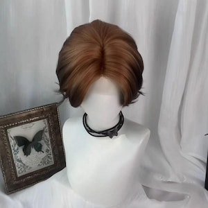 May include: A brown and auburn wig with a short, layered style is displayed on a white mannequin head. The wig has a natural-looking hairline and is styled with soft waves. A black choker necklace with a butterfly pendant is around the mannequin's neck.