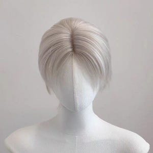 May include: A short, light gray wig with a center part, styled on a white mannequin head. The wig has a natural-looking hairline and a slightly textured appearance, suggesting a realistic hair fiber. The mannequin's shoulders are also visible.