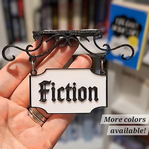 May include: A black and white "Fiction" sign with decorative scrollwork. The sign is held up by a decorative black metal bracket. The text "More colors available!" is below the sign.