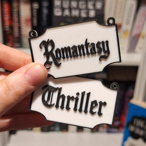 May include: Two white rectangular signs with black trim and text. The top sign reads "Romantasy" in a decorative font. The bottom sign reads "Thriller" in a similar font. The signs have small holes at the top for hanging.