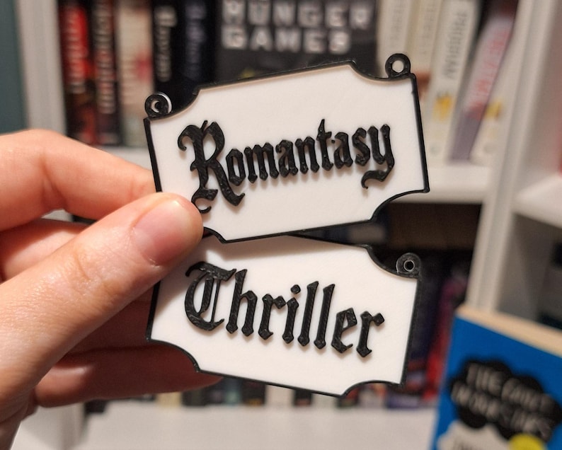May include: Two white rectangular signs with black trim and text. The top sign reads "Romantasy" in a decorative font. The bottom sign reads "Thriller" in a similar font. The signs have small holes at the top for hanging.