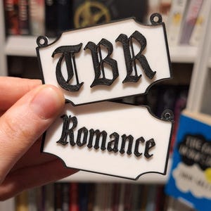 May include: Two white rectangular book markers with black lettering. The top marker reads "TBR" and the bottom marker reads "Romance". The markers have decorative edges and small holes at the top for hanging. The background shows a bookshelf with books.
