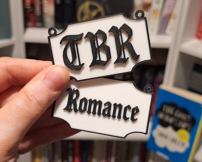 May include: Two white rectangular book markers with black lettering. The top marker reads "TBR" and the bottom marker reads "Romance". The markers have decorative edges and small holes at the top for hanging. The background shows a bookshelf with books.