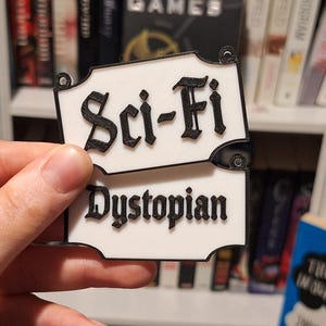 May include: White and black Sci-Fi Dystopian book label. The text is in a gothic font. The label is held in a hand in front of a bookshelf filled with books.