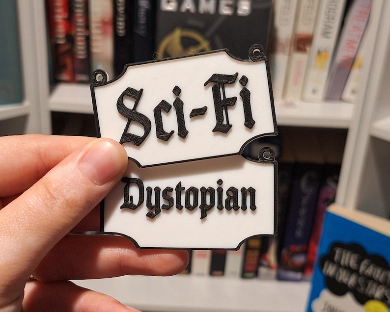 May include: White and black Sci-Fi Dystopian book label. The text is in a gothic font. The label is held in a hand in front of a bookshelf filled with books.