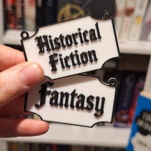 May include: Two white rectangular book labels with black trim and text. The top label reads "Historical Fiction" and the bottom label reads "Fantasy". The labels have a decorative shape and small holes for hanging.