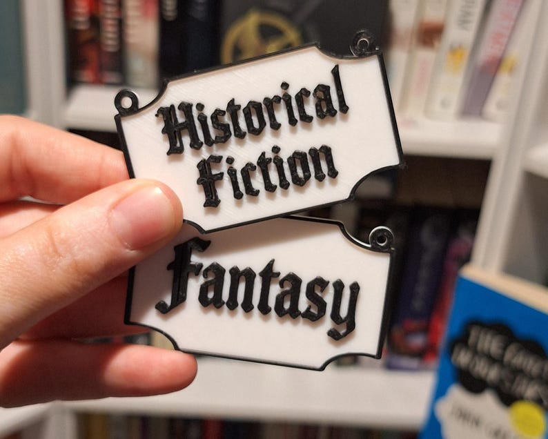 May include: Two white rectangular book labels with black trim and text. The top label reads "Historical Fiction" and the bottom label reads "Fantasy". The labels have a decorative shape and small holes for hanging.