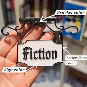 May include: A black and white sign that reads "Fiction" in a decorative font. The sign is suspended from a black bracket with ornate scrollwork. The sign has a white background and a black border.