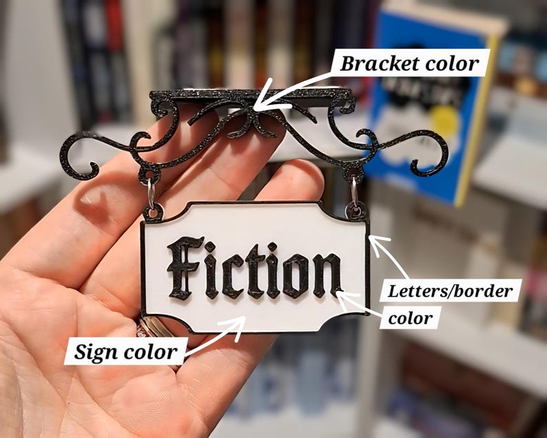 May include: A black and white sign that reads "Fiction" in a decorative font. The sign is suspended from a black bracket with ornate scrollwork. The sign has a white background and a black border.