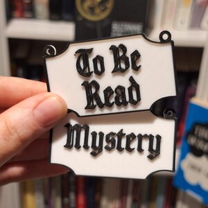 May include: Two white, decorative signs with black text that read "To Be Read" and "Mystery". The signs are held in a hand, with a bookshelf in the background. The signs have a decorative shape with small holes at the top.