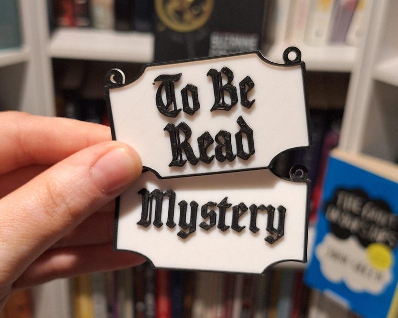 May include: Two white, decorative signs with black text that read "To Be Read" and "Mystery". The signs are held in a hand, with a bookshelf in the background. The signs have a decorative shape with small holes at the top.
