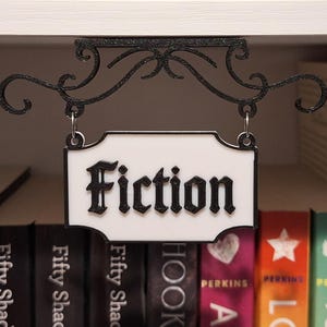 May include: A black metal sign with the word "Fiction" in a decorative font hangs above a shelf of books. The sign is white with a black border and is suspended by a decorative scrolled bracket.