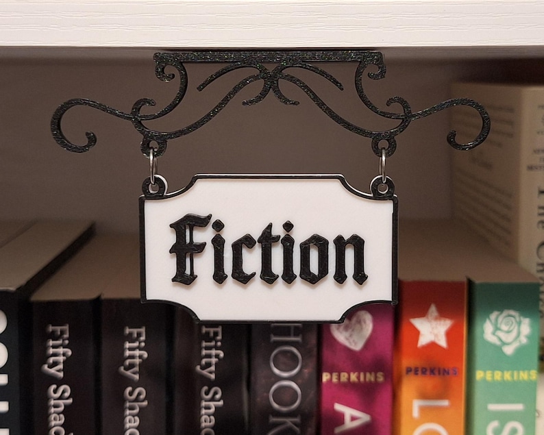 May include: A black metal sign with the word "Fiction" in a decorative font hangs above a shelf of books. The sign is white with a black border and is suspended by a decorative scrolled bracket.