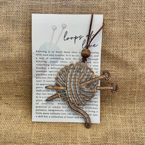 May include: Wooden pendant featuring a stylized yarn ball with knitting needles. The pendant is attached to a leather cord and a wooden bead. The card behind the pendant has the text "loops of love" and text about knitting.