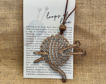 May include: Wooden pendant featuring a stylized yarn ball with knitting needles. The pendant is attached to a leather cord and a wooden bead. The card behind the pendant has the text "loops of love" and text about knitting.
