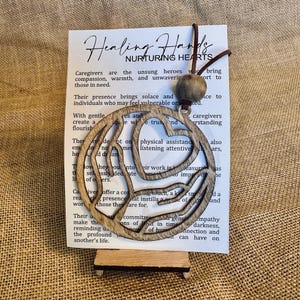 May include: Wooden cutout of two hands forming a heart shape with the text "Healing Hands Nurturing Hearts" on a white card. The text on the card reads, "Caregivers are the unsung heroes who bring compassion, warmth, and unwavering support to those in need."