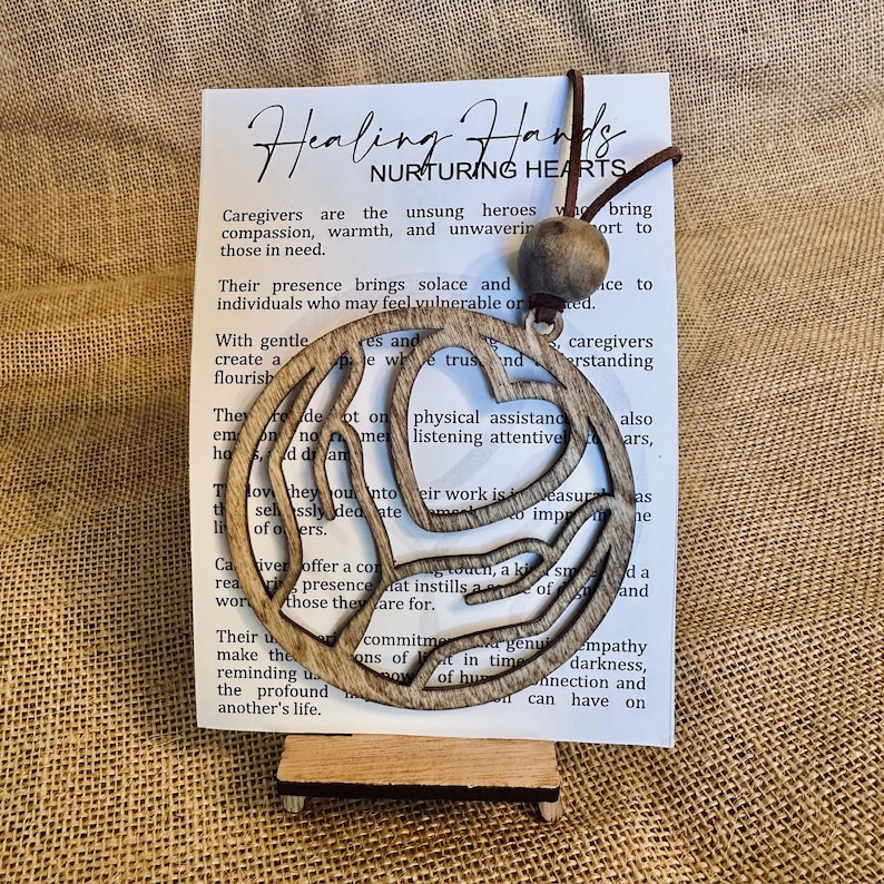 May include: Wooden cutout of two hands forming a heart shape with the text "Healing Hands Nurturing Hearts" on a white card. The text on the card reads, "Caregivers are the unsung heroes who bring compassion, warmth, and unwavering support to those in need."