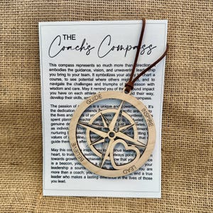 May include: Wooden compass ornament with the text "The Coach's Compass" and the words "Guide", "Believe", "Lead", and "Inspire". The compass is attached to a white card with text and a brown leather cord.