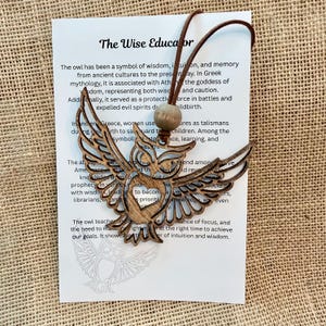 May include: A wooden owl ornament with intricate cut-out details, hanging from a brown cord. The ornament is displayed with a card that reads "The Wise Educator" and text about the owl's symbolism.