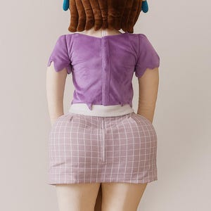 May include: A plush doll with a brown wig, blue earrings, and a purple top and skirt. The skirt has a pink and white grid pattern. The doll is wearing light tan leggings and purple boots. The doll is facing away from the camera.