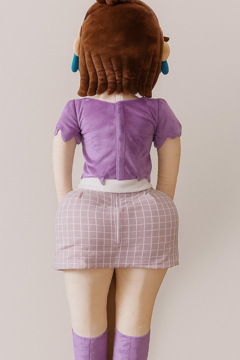 May include: A plush doll with a brown wig, blue earrings, and a purple top and skirt. The skirt has a pink and white grid pattern. The doll is wearing light tan leggings and purple boots. The doll is facing away from the camera.