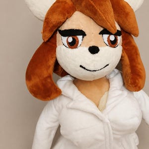 Puede incluir: Un peluche que representa a un personaje antropomórfico con cabello castaño, ojos grandes de color marrón y cejas oscuras. El personaje tiene un hocico color crema, una pequeña nariz negra y una fina sonrisa negra. Viste una bata blanca con cuello.