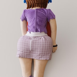 May include: A plush doll of a character, viewed from the back. The doll has brown hair, a purple top, a plaid skirt, and purple boots. A red cape is visible. The doll is leaning against a white wall.