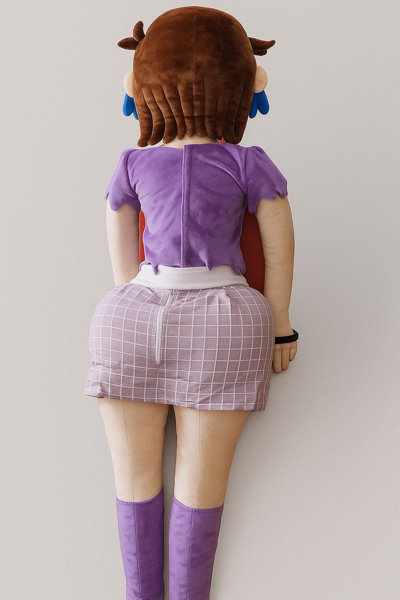 May include: A plush doll of a character, viewed from the back. The doll has brown hair, a purple top, a plaid skirt, and purple boots. A red cape is visible. The doll is leaning against a white wall.