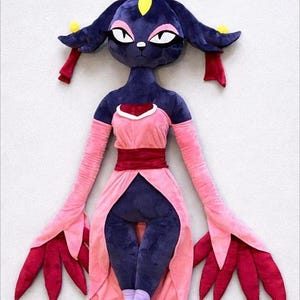May include: A plush doll in the shape of a character with a dark blue head and body, wearing a pink dress with a red belt. The doll has large, expressive eyes and long, flowing arms with red petal-like details at the ends.