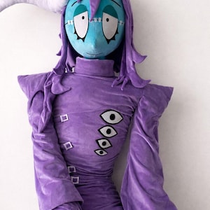May include: A plush doll with a blue face, purple hair, and a purple outfit. The doll has stitched features and multiple eye designs on the chest. A white horn-like shape extends from the head.