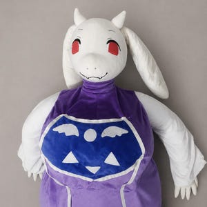 Puede incluir: Una muñeca de peluche de un personaje con cabeza blanca, ojos rojos y orejas largas. La muñeca lleva un vestido morado con un emblema azul en el pecho. La muñeca tiene brazos y manos blancos.