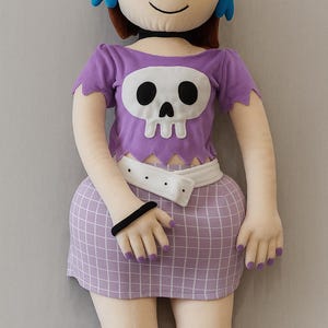 May include: A plush doll of a cartoon character with brown hair, purple eyeshadow, and blue earrings. The doll is dressed in a purple crop top with a skull design, a white belt, and a checkered skirt. Purple socks and a black bracelet complete the outfit.