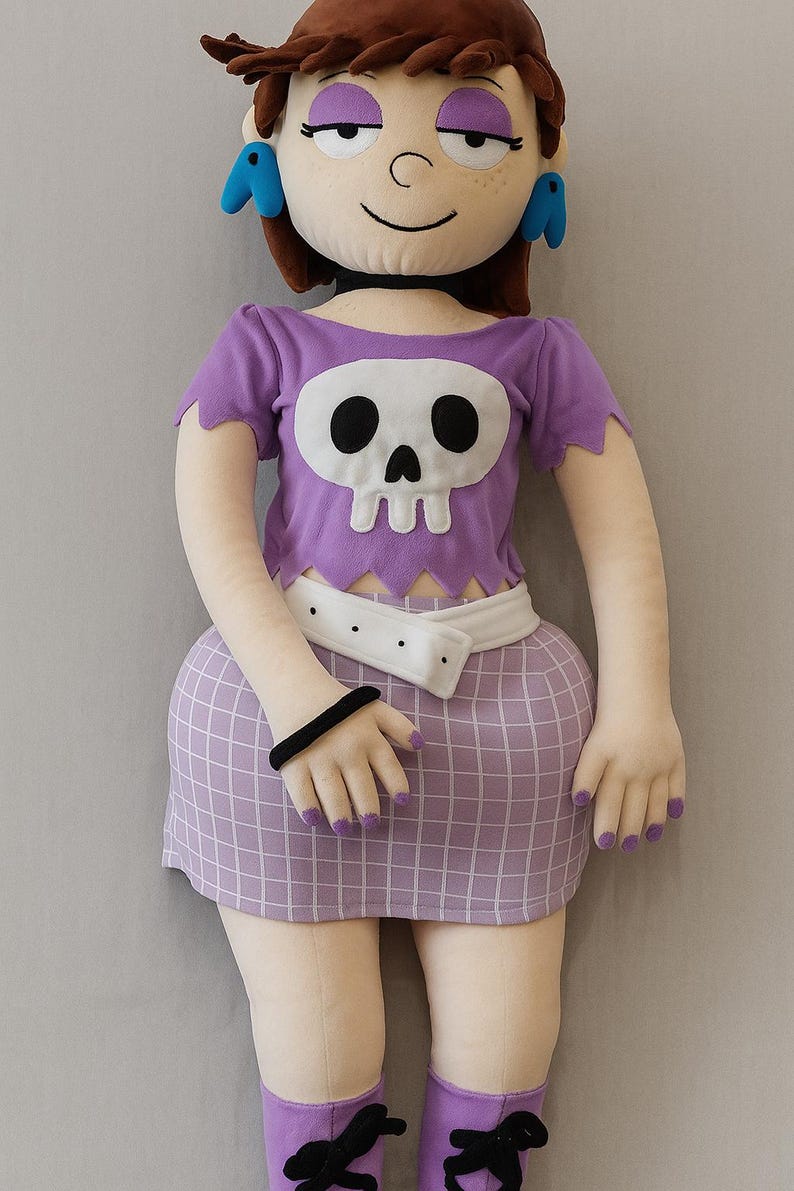 May include: A plush doll of a cartoon character with brown hair, purple eyeshadow, and blue earrings. The doll is dressed in a purple crop top with a skull design, a white belt, and a checkered skirt. Purple socks and a black bracelet complete the outfit.