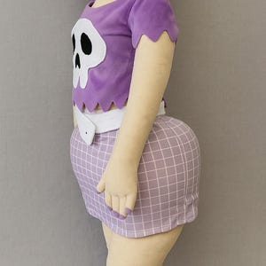 May include: A plush doll dressed in a purple top featuring a white skull motif and a coordinating checkered skirt. The doll is wearing a white belt and is viewed from the side. The doll's arms and legs are visible.