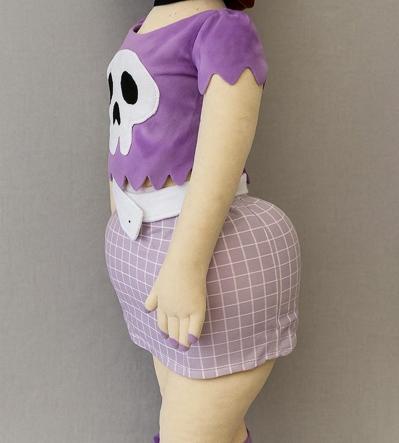 May include: A plush doll dressed in a purple top featuring a white skull motif and a coordinating checkered skirt. The doll is wearing a white belt and is viewed from the side. The doll's arms and legs are visible.