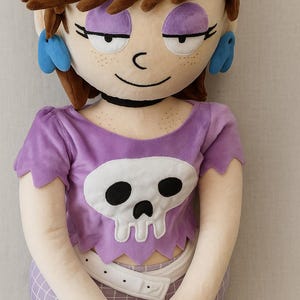May include: A plush doll of a cartoon character with brown hair, purple eyeshadow, and blue earrings. The doll wears a purple shirt with a white skull design and a matching skirt. A black bracelet and choker complete the look.