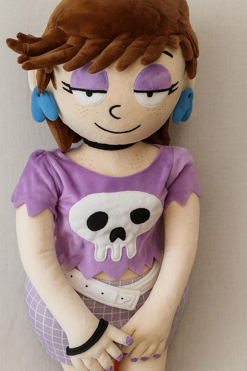 May include: A plush doll of a cartoon character with brown hair, purple eyeshadow, and blue earrings. The doll wears a purple shirt with a white skull design and a matching skirt. A black bracelet and choker complete the look.