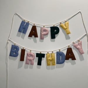 May include: A fabric banner spelling "HAPPY BIRTHDAY". The letters are in blue, brown, pink, green, and yellow, attached to a string with wooden pegs. A cheerful decoration for a birthday celebration.