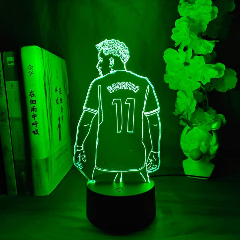 May include: A green-glowing LED lamp featuring a soccer player in a jersey with the name "RODRYGO" and the number "11" on the back. The lamp is on a black base, with books and a decorative plant in the background.