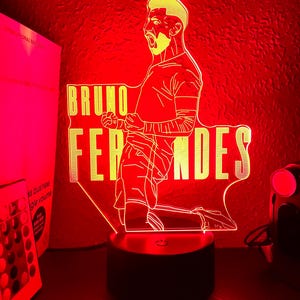 Op de afbeelding: Een rood verlichte LED-lamp met een gestileerde illustratie van een voetballer met de naam "BRUNO FERNANDES" in het geel. De lamp staat op een zwarte voet, met een rode achtergrond.