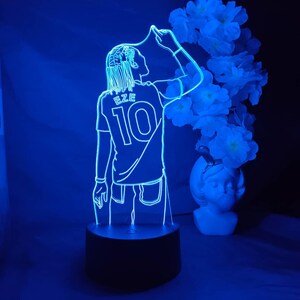 May include: A blue LED lamp depicting a footballer in a jersey with the name "EZE" and the number "10". The lamp sits on a black base, with a vase of white flowers in the background.