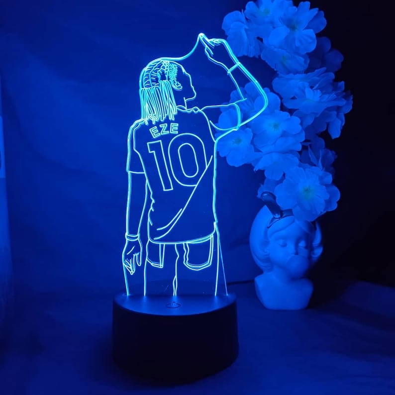May include: A blue LED lamp depicting a footballer in a jersey with the name "EZE" and the number "10". The lamp sits on a black base, with a vase of white flowers in the background.