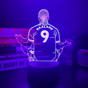May include: A purple-lit LED lamp featuring a stylized outline of a soccer player's jersey with the name "HAALAND" and the number "9" on the back. The lamp is on a black base, with books in the background.
