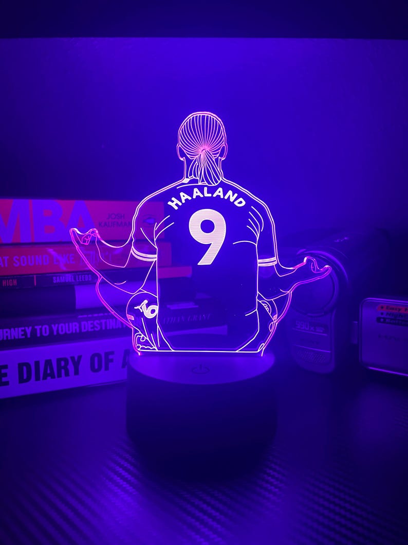 May include: A purple-lit LED lamp featuring a stylized outline of a soccer player's jersey with the name "HAALAND" and the number "9" on the back. The lamp is on a black base, with books in the background.