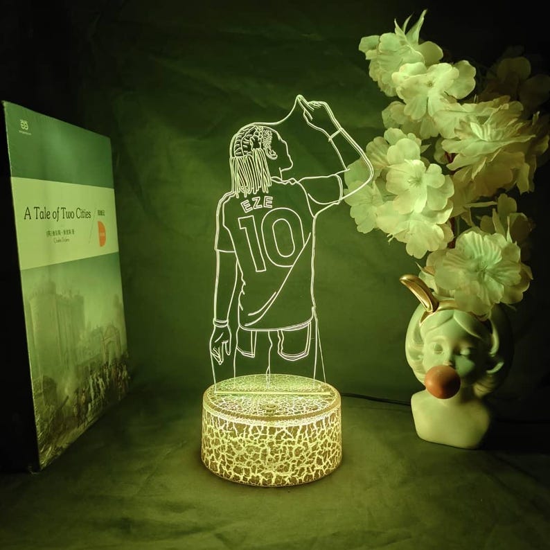 May include: An LED lamp with a white outline of a person in a jersey with "EZE" and "10". The lamp sits on a round base with a cracked pattern. A book and a vase with flowers are in the background. The lamp is a decorative item.