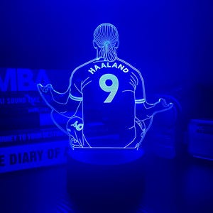 May include: A blue-lit LED lamp featuring a soccer player's jersey with the name "HAALAND" and the number "9" on the back. The lamp is designed in a minimalist style, with the player's silhouette and jersey details outlined in white.