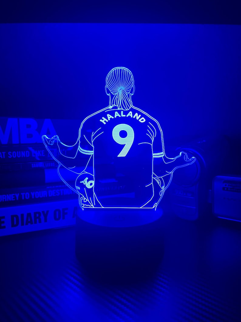 May include: A blue-lit LED lamp featuring a soccer player's jersey with the name "HAALAND" and the number "9" on the back. The lamp is designed in a minimalist style, with the player's silhouette and jersey details outlined in white.