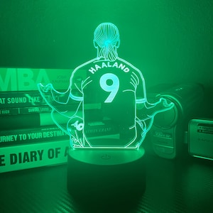 May include: A green-lit LED lamp featuring a soccer player in a meditative pose. The player's jersey displays "HAALAND" and the number "9". The lamp sits on a black base, with books and electronics in the background.